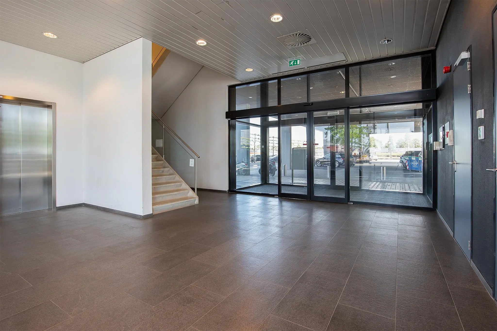 Ingangshal van een modern kantoorgebouw aan de Waalhaven O.Z. met glazen schuifdeuren, trap, lift en grijze vloertegels.